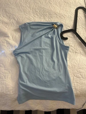 Ljusblå oneshoulder topp - Snygg ljusblå oneshoulder topp med dekorativ detalj vid axeln. Toppen är ärmlös och har en tight passform, perfekt för att lyfta din outfit med en unik twist.