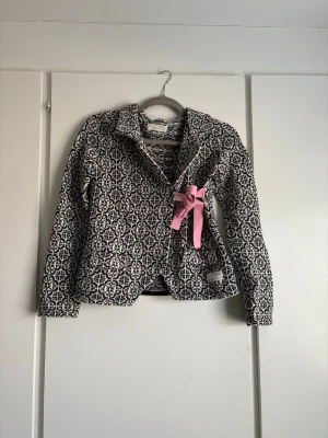 Mönstrad svartvit jacka/kofta med rosa rosett - Unik svartvit jacka/kodta med grafiskt mönster och en söt rosa rosett framtill. Jackan/koftan har lång ärm, knappar och figurnära passform. Perfekt för dig som vill sticka ut med en cool detalj. Tillverkad i mjukt material som känns bekvämt hela dagen. Pris kan diskuteras💕