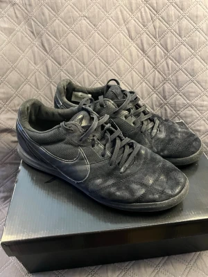 Svarta Nike fotbollsskor - Säljer ett par helsvarta fotbollsskor från Nike med klassisk låg profil och snörning. Skorna har en stilren design med diskret Nike-logga på sidan och ovandel i syntetmaterial och mesh för bra ventilation. Perfekta för dig som vill ha en clean look på planen. Sliten sula 