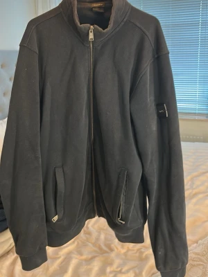 Svart ziptröja  - Snygg svart ziptröja från Oscar Jacobson med hög krage och dragkedja framtill. Tröjan har två fickor med dragkedja och ribbade muddar vid ärmslut och nederkant. Perfekt för en stilren och avslappnad look.