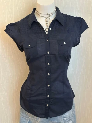 Mörkblå kortärmad skjorta med knappar - Snygg mörkblå skjorta med korta ärmar och vita knappar framtill. Skjortan har två bröstfickor med lock och en figurnära passform. Materialet ser ut att vara bomull eller en bomullsblandning, perfekt för en stilren look.