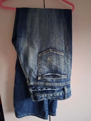 Blå jeans med broderi och fågelmotiv - Säljer ett par blå jeans med snyggt slitna detaljer och broderat fågelmotiv på bakfickan samt broderier längs benet. Jeansen har klassisk femficksmodell och är tillverkade i denim. Perfekta för dig som vill ha något unikt och trendigt i garderoben.