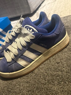 Adidas Campus blå sneakers - Säljer ett par klassiska Adidas Campus sneakers i blå mocka med vita detaljer och breda vita skosnören. Kommer med 3 olika slags snören, skoboxen.. Inga defekter, bara smutsiga 