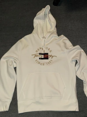 Vit hoodie från Tommy Hilfiger - Snygg vit hoodie från Tommy Hilfiger med broderad logga och texten 'New York Since 1985' i guld. Klassisk känguruficka framtill och justerbar huva med snören. Perfekt för en avslappnad och trendig stil.