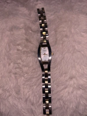 Fossil silver och guld armbandsur - Stilren klocka från Fossil med smalt armband i silver och guldton. Urtavlan är vit med minimalistiska detaljer och guldfärgade visare. Klockan har en elegant, avlång form och länkar i metall som ger en klassisk look.