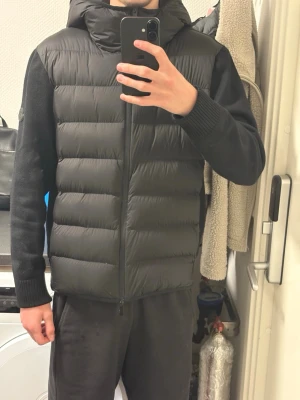 Svart Moncler pufferjacka med huva - Svart pufferjacka från Moncler med stickade ärmar och huva. Jackan har quiltad framsida i syntetmaterial och dragkedja hela vägen. Moncler-logga på ärmen. Perfekt för dig som gillar stilren och sportig look.