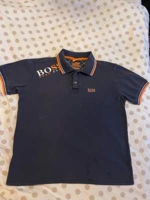 Marinblå pikétröja från BOSS - Snygg marinblå pikétröja från BOSS med orange och vita detaljer på krage och ärmslut. Klassisk pikéknäppning framtill och logga på bröstet samt axeln. Perfekt för en clean och sportig look.