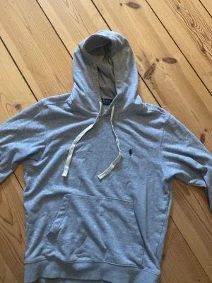 Grå hoodie från Polo Ralph Lauren - Säljer en grå hoodie från Polo Ralph Lauren med klassisk huva och vita snören. Tröjan har en känguruficka framtill och den ikoniska Polo-loggan broderad på bröstet. Perfekt för chill dagar och enkel att matcha med jeans eller joggers.