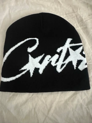 Cortiez mössa  - Säljer en svart beanie med stor vit text och stjärndetaljer i grafisk stil. Mössan har en klassisk form och är stickad i mjukt material, perfekt för kalla dagar. Snygg streetwear-vibe och passar till de flesta outfits.