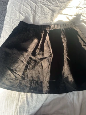 Svart kort kjol med resårmidja - Säljer en svart kort kjol med resår i midjan. Kjolen har en lätt glansig yta och är gjord i ett tunt, lite prassligt material som ger en avslappnad look. Perfekt att styla med en crop top eller hoodie för en chill vibe. Helt ny oanvänd 