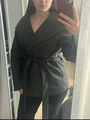 Mörkgrå omlottkappa med knytskärp - Snygg mörkgrå omlottkappa med bred krage och knytskärp i midjan. Kappan har en avslappnad passform och är tillverkad i ett mjukt tyg som ger en stilren look. Perfekt för lager på lager och funkar till många outfits.