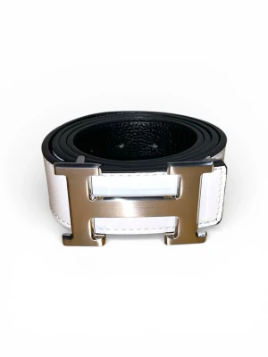 Vitt skärp med H-spänne - Säljer ett stilrent vitt skärp med det ikoniska H-formade metallspännet. Skärpet är tillverkat i skinn och har en klassisk design som lyfter vilken outfit som helst. Insidan är svart och spännet har en elegant silverfinish.