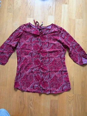 Röd paisleymönstrad blus med knyt - Snygg röd blus med paisleymönster i vitt och svart. Blusen har trekvartsärm och en dekorativ volang vid halsringningen samt knytband i nacken. Materialet känns lätt och luftigt, perfekt för en trendig look.