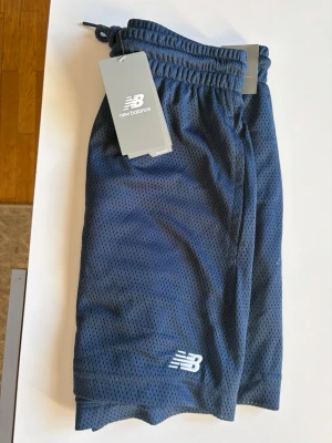 Blå meshshorts från New Balance - Snygga marinblå shorts från New Balance i meshmaterial. De har elastisk midja med snörning och en liten vit logga på benet. Perfekta för träning eller chill dagar. Luftiga och bekväma med sportig vibe.