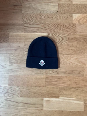 Svart Moncler mössa med logga - Säljer en svart ribbstickad mössa från Moncler med uppvikt kant och den klassiska Moncler-loggan framtill. Mössan har en enkel och clean design som passar till det mesta. Perfekt för dig som vill ha en stilren accessoar med exklusiv känsla.