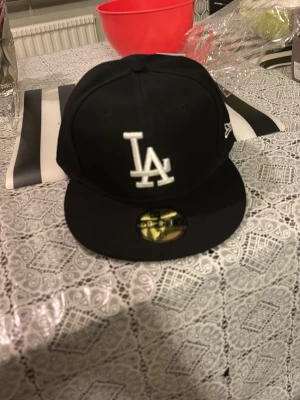 Svart LA-keps från New Era - Snygg svart keps från New Era med LA-logga broderad i vitt framtill. Klassisk flat peak och New Era-logga på sidan. Perfekt accessoar för en sportig och trendig stil.