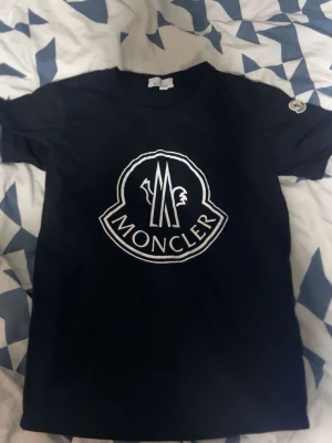 Svart Moncler t-shirt med stor logga - Snygg svart t-shirt från Moncler med stor vit logga på bröstet och Moncler-märke på ärmen. Klassisk rund hals och korta ärmar. Perfekt för dig som gillar stilrena och exklusiva plagg. Det står M på tröjan men de är verkligen passar de xs-s 