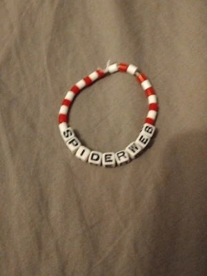 Rött och vitt pärlarmband SPIDERWEB - Armband med röda och vita plastpärlor samt vita bokstavspärlor som stavar 'SPIDERWEB'. Armbandet har en enkel, rund form och är elastiskt för att passa de flesta handleder. Perfekt för dig som gillar personliga och färgglada accessoarer.