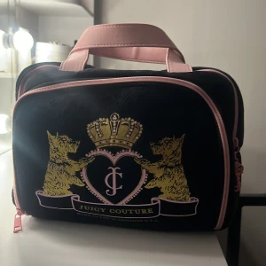 Juicy Couture svart och rosa necessär - Snygg necessär från Juicy Couture i svart sammet med rosa detaljer och handtag. Framsidan har ett broderat emblem med två hundar, krona och hjärta. Insidan är ljusrosa med meshfickor för smart förvaring. Perfekt för smink eller småprylar.