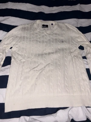 Vit kabelstickad tröja från GANT - Kabelstickad vit tröja från GANT i premium bomull. Klassisk rund halsringning och diskret GANT-logga broderad på bröstet. Perfekt för dig som gillar stilrena och tidlösa plagg med snygg struktur.