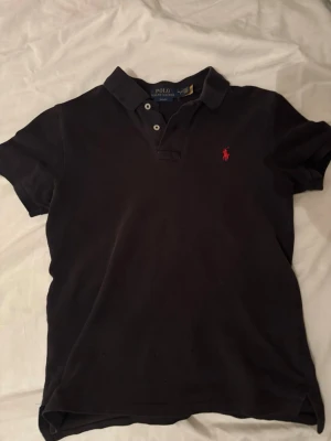 Svart piké från Polo Ralph Lauren - Klassisk svart pikétröja från Polo Ralph Lauren med slim fit-passform. Tröjan har korta ärmar, krage och knappar framtill samt den ikoniska röda logotypen broderad på bröstet. Perfekt för en stilren och avslappnad look.