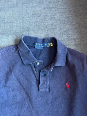 Marinblå piké från Ralph Lauren - Klassisk marinblå pikétröja från Ralph Lauren med röd broderad logga på bröstet. Slim fit-modell med krage och två knappar framtill. Tillverkad i mjuk bomull som känns skön mot huden. Perfekt för en clean och stilren look. Kan vara lite solbränd men inget jag tänkt på! Pris kan diskuteras