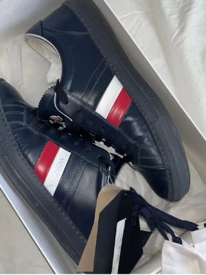 Moncler sneakers i marinblått skinn - Snygga sneakers från Moncler i marinblått skinn med svarta skosnören och en ikonisk röd och vit rand på sidan. Klassisk låg modell med svart sula och diskret Moncler-logga på sidan. Perfekta för dig som gillar stilrena och exklusiva detaljer.