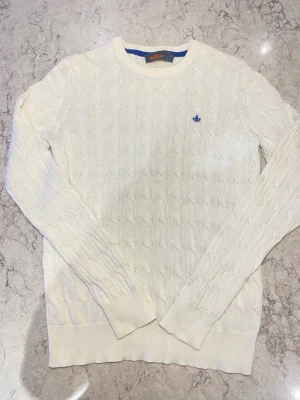 Vit kabelstickad tröja Morris - Kabelstickad vit tröja från Morris med rund halsringning och diskret blå logga broderad på bröstet. Tröjan har långa ärmar och ribbade muddar vid ärmslut och nederkant. Perfekt för dig som gillar klassisk och stilren design.