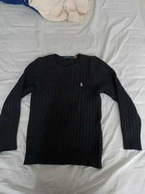 Svart kabelstickad tröja Polo Ralph Lauren - Kabelstickad svart tröja från Polo Ralph Lauren med rund halsringning och den klassiska broderade loggan i vitt på bröstet. Långa ärmar och ribbade muddar vid ärmslut och nederkant. Perfekt för en stilren och enkel look.