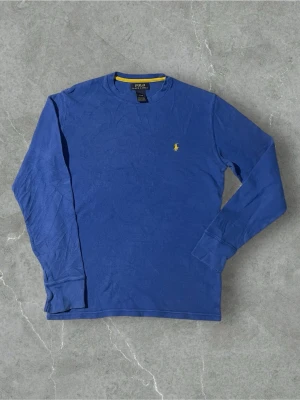 Blå långärmad tröja Polo Ralph Lauren | S - Blå långärmad tröja från Polo Ralph Lauren med rund halsringning och gul broderad logga på bröstet. Tröjan har en diskret textur i materialet och en gul detalj på insidan av kragen. Perfekt för en avslappnad och stilren look.