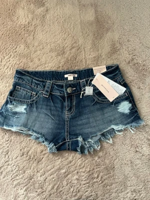 Blå jeansshorts från Gina tricot 14+ - Säljer ett par blå jeansshorts från Gina tricot 14+ med slitna detaljer och fransig kant. Shortsen har fem fickor, bälteshällor och klassisk knappgylf. Perfekta för sommaren och har en cool, avslappnad vibe. Stora i storleken.