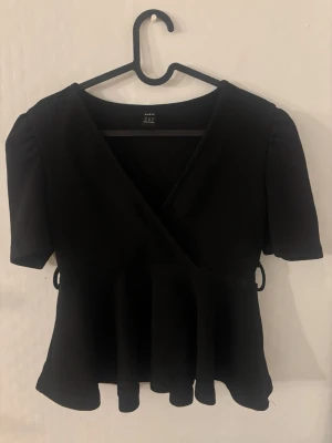 Svart omlottopp med volang - Snygg svart omlottopp från shein med korta puffärmar och volang nedtill. Toppen har en markerad midja och omlottdesign som ger en feminin siluett. Perfekt att styla med jeans eller kjol för en trendig look.