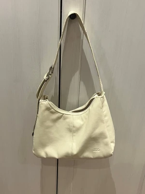 Krämvit axelväska från JW PEI - Stilren axelväska i krämvitt veganskt läder från JW PEI. Väskan har en justerbar axelrem och minimalistisk design med diskret logga framtill. Perfekt för dig som gillar enkel och trendig look.