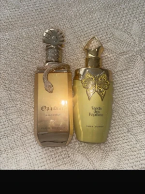 Paris Corner parfym duo guld - Två exklusiva parfymer från Paris Corner: Ophidian Mango Bliss i en guldskimrande flaska med ormdekor och Jardin des Papillons i en elegant guldfärgad flaska med fjärilsmotiv. Båda flaskorna har unika, detaljerade lock och lyxig design.