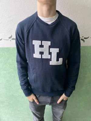 Mörkblå sweatshirt med HL-tryck - Mörkblå sweatshirt med stora vita bokstäver 'HL' på bröstet. Tröjan har rund halsringning, ribbade muddar vid ärmslut och nederkant samt en avslappnad passform. Perfekt för en casual och sportig stil.