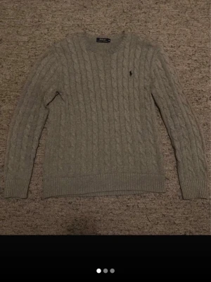 Ralph Lauren tröja - Säljer en grå kabelstickad tröja från Polo Ralph Lauren med rund halsringning och den klassiska svarta logotypen broderad på bröstet. Tröjan har långa ärmar och ribbade muddar vid ärmslut och nederkant. Perfekt för lager på lager under kyliga dagar. Bara att skriva vid frågor!