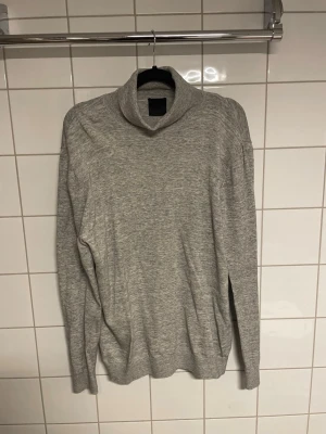 Ljusgrå polotröja i stickat material - Säljer en stilren ljusgrå stickad polotröja med lång ärm. Tröjan har en hög krage och är tillverkad i ett mjukt, tunt material som passar perfekt till lager-på-lager. Enkel och clean design utan synliga loggor.