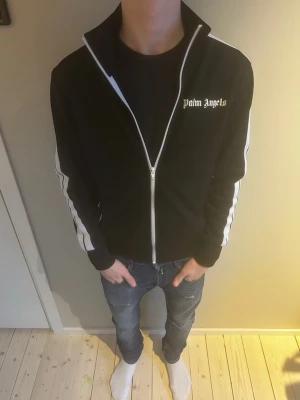 Palm Angels svart ziptröja - Svart ziptröja från Palm Angels med vit logga på bröstet och vita ränder längs ärmarna. Hög krage och dragkedja framtill ger en sportig vibe. Perfekt för dig som gillar streetwear och vill sticka ut med en clean look. Själv klart äkta, inga kopior! 