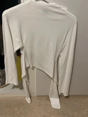 Vit tröja man knyter i ryggen  - Säljer en vit omlottopp med långa ärmar och långa knytband i midjan. Toppen har en asymmetrisk nederkant och är tillverkad i ett mjukt, stretchigt material som känns skönt mot huden. Perfekt för dig som gillar trendiga och stilrena plagg.