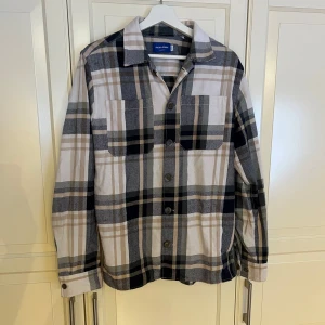 Rutig Jack & Jones skjortjacka / overshirt - Skjortjacka/overshirt i rutigt mönster från Jack & JonesSnygg och trendig skjortjacka/overshirt från Jack & Jones i ett stilrent rutigt mönster i vitt, beige, blått och svart. Perfekt som lager-på-lager-plagg under vår och höst, eller som lätt jacka svalare sommarkvällar.  Plagget är i gott skick och har endast använts ett fåtal gånger. Inga fläckar eller hål. Nypris: 499,95 kr. 