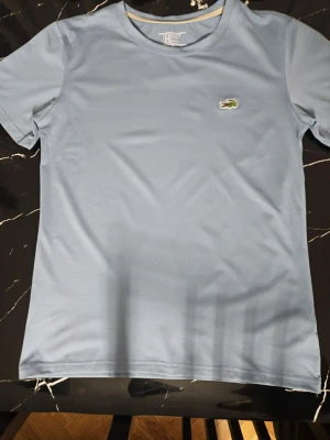 Ljusblå t-shirt från Lacoste - Snygg ljusblå t-shirt från Lacoste med klassisk rund hals och det ikoniska krokodilmärket broderat på bröstet. Perfekt för en clean och stilren look. Storlek XS