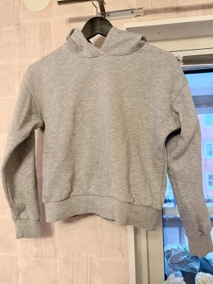 Grå hoodie från KappAhl - En enkel och stilren grå hoodie från KappAhl i mjukt sweatshirtmaterial. Hoodien har ribbade muddar vid ärmslut och nederkant samt en klassisk huva utan snören. 