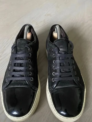 Svarta sneakers med lackdetaljer - Snygga svarta sneakers med glansiga lackdetaljer på tån och klassisk vit sula. Skorna har svarta skosnören och metallhål för snörningen. Perfekta för dig som vill ha en stilren men ändå unik look. Passar till både jeans och chinos.
