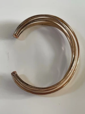 Guldigt öppet armband i metall - Snyggt armband i guldfärgad metall med öppen design. Armbandet har tre parallella band som ger en modern och stilren look. Perfekt accessoar för att lyfta din outfit med en touch av guld.