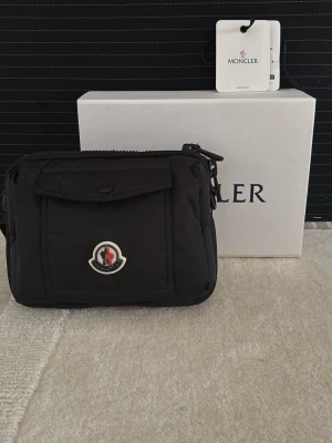 Svart Moncler handväska nylon - Snygg svart handväska från Moncler i slitstark nylon med klassisk logotyp framtill. Väskan har en framficka med lock och knapp, justerbar axelrem och stilren design. Perfekt för dig som gillar exklusiva accessoarer med sportig touch. Fick denna i present för längesen och har aldrig använt den sen dess då jag inte tycker jag passar i den! Den är 100% äkta och vi säljer inte fake här på Sverigesresell så de behöver ni inte oroa er för! Box tillkommer