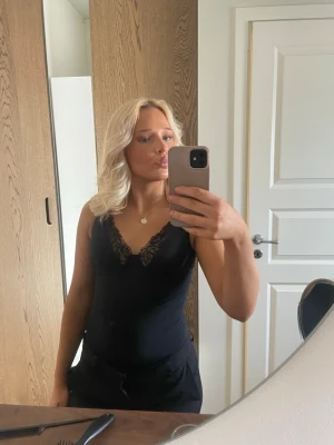 Svart spetsbody med v-ringning - Säljer en svart body med v-ringning och fina spetsdetaljer vid urringningen. Bodyn har smala axelband och är figurnära, vilket ger en snygg siluett. 