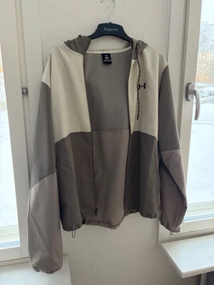 Beige och grå vindjacka från Under Armour - Snygg vindjacka från Under Armour i beige och grå färgblock. Jackan har huva, dragkedja framtill och justerbar nederkant med dragsko. Tillverkad i lätt och tåligt material, perfekt för blåsiga dagar. Diskret logga på bröstet och meshfoder för extra komfort.