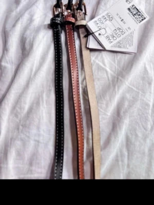 Set med smala bälten i skinn från hm oanvända  - Säljer ett set med tre smala bälten i skinn. Ett är svart med vita sömmar, ett är brunt med vita sömmar och det tredje är beige. Alla har silverfärgade spännen och klassisk design som passar till jeans eller klänning. 