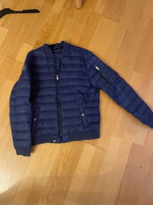 Blå pufferjacka från Polo Ralph Lauren - Snygg mörkblå pufferjacka från Polo Ralph Lauren med quiltad design och ribbade muddar. Jackan har dragkedja framtill, fickor med knappar och en patch på ärmen. Perfekt för kalla dagar och riktigt stilren look.