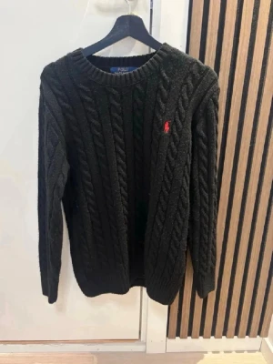 Svart kabelstickad tröja Polo Ralph Lauren - Kabelstickad svart tröja från Polo Ralph Lauren med rund halsringning och röd broderad logga på bröstet. Tröjan har långa ärmar och ribbade muddar vid ärmslut och nederkant. Perfekt för dig som gillar klassisk och stilren look.Kom dm för eventuella frågor 🧐pris kan alltid diskuteras 😊